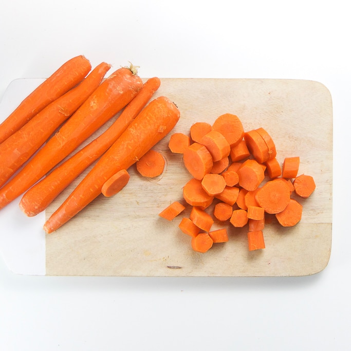 a wooden cutting board sitting on a white background with a bunch of peeled carrots sitting on the left hand side with some chopped carrots sitting on the other.