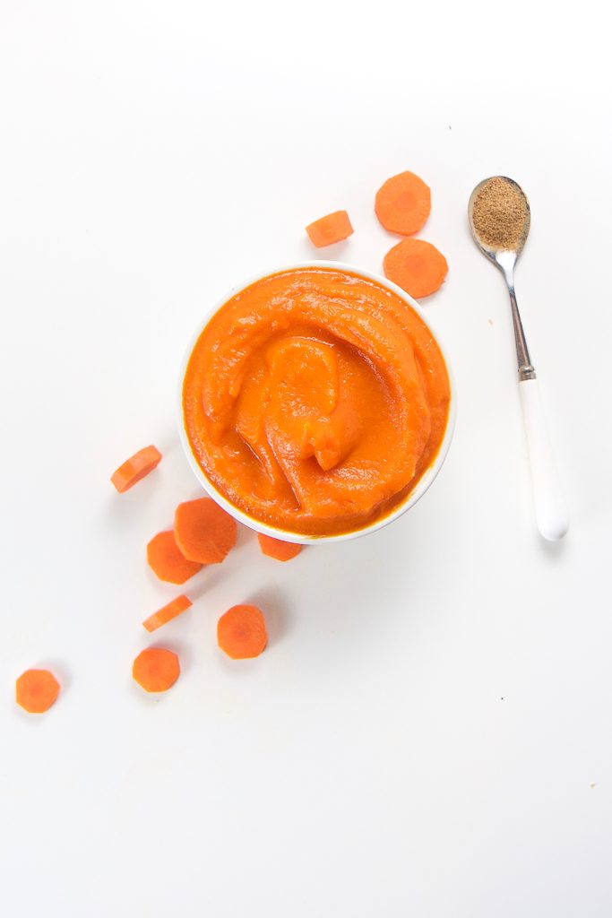 small white bowl sitting on a white background with a bring carrot baby food puree inside with a few chopped carrot coins on the surface and a white and silver spoon with nutmeg inside.