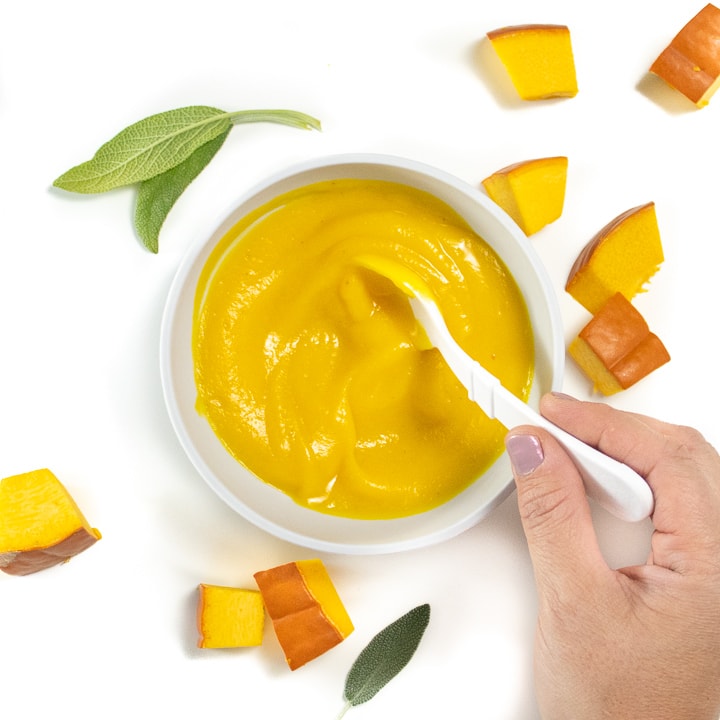 Gray bowl with pumpkin puree with chunks of pumpkin and sage around it. Hand is holding a white spoon with pure on it.