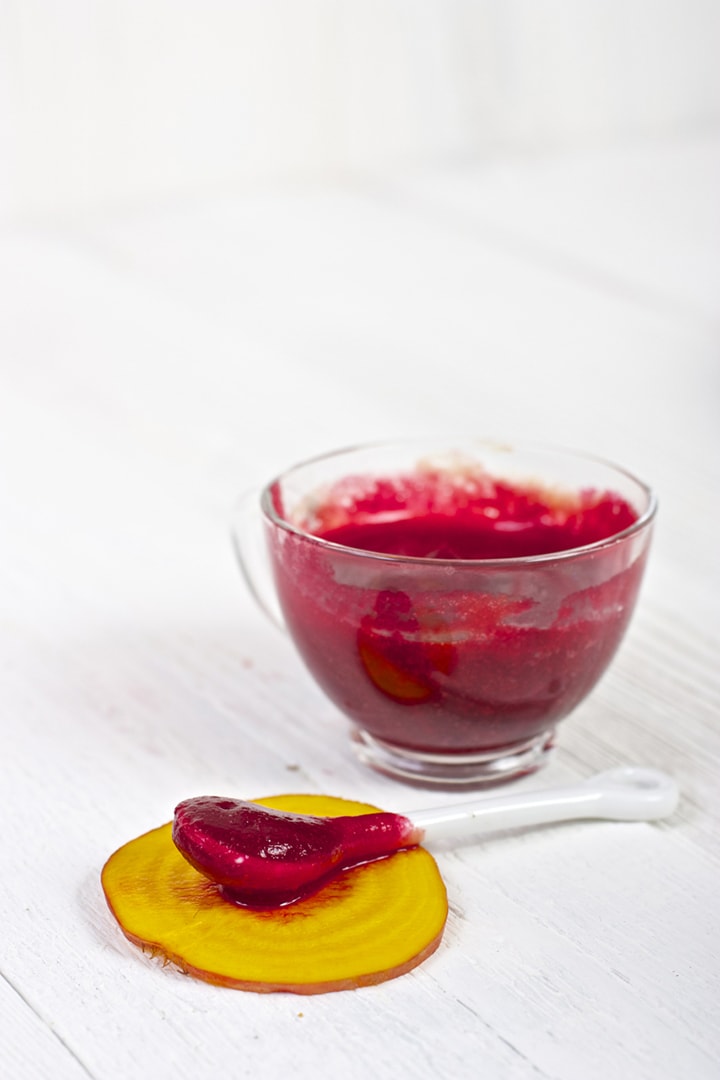 clear jar filled with red beet puree with a spoon sitting in front of it resting on a piece of golden beet.