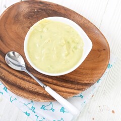 white bowl sitting on an oval wooden plate with a silver and white spoon resting on top. The small white bowl is filled with an egg and avocado baby food puree.