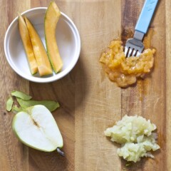 On a wooden cutting board we have one side slices of pear, cantaloupe and avocado and the other side is them smashed with a fork.