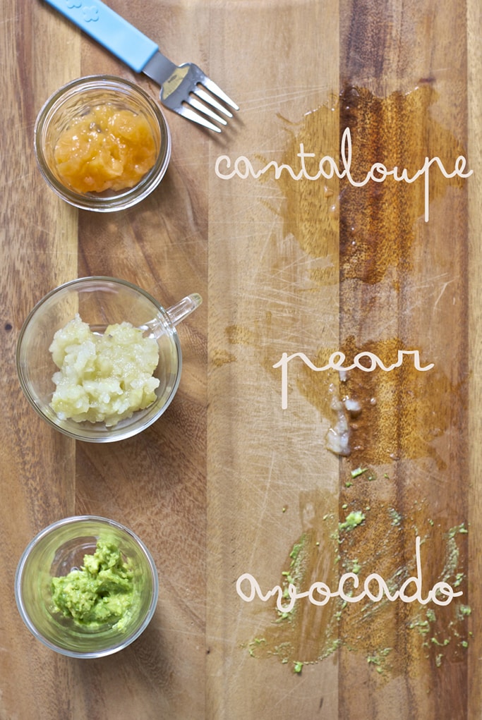 On a wooden cutting board is 3 small glass jars filled with the smashed baby food - pear, avocado and cantaloup with their names next to them. 