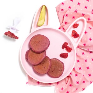 Pink bunny plate for baby or toddler filled with pink pancakes, raspberries and avocado sitting on a pink napkin with a white fork for baby to use.