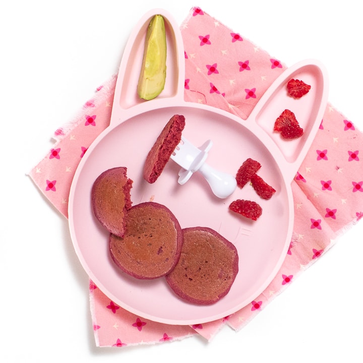 Pink bunny plate for baby or toddler filled with pink pancakes, raspberries and avocado sitting on a pink napkin with a white fork for baby to use.