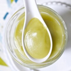 Image is of a white spoon resting on a clear jar which are both filled with a baby food puree.