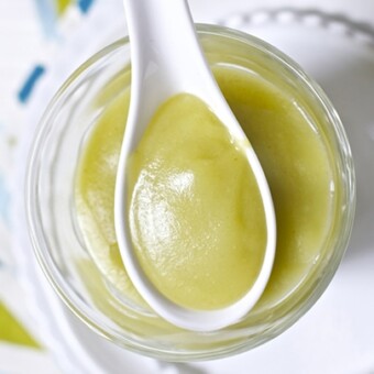 Image is of a white spoon resting on a clear jar which are both filled with a baby food puree.