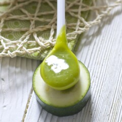 A small deep white spoon is filled with a green zucchini baby food puree and is sitting on top of a small round slice of zucchini. All of this is sitting on a white wood surface.