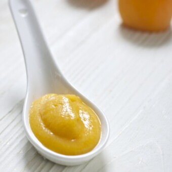 white spoon filled with a thick and creamy apricot and pear baby food puree. Spoon is sitting on a white wooden board and there are some fresh apricots in the background.