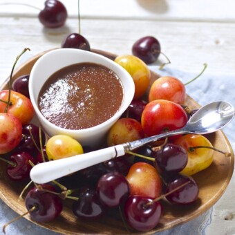 small white bowl sitting on a wooden plate that is filled with whole cherries. In the bowl is a thick red cherry baby food puree.