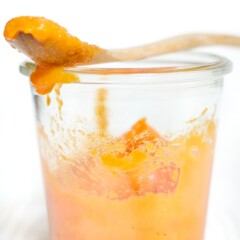 Peach and carrot baby food puree in a clear jar with a wooden spoon resting on top.