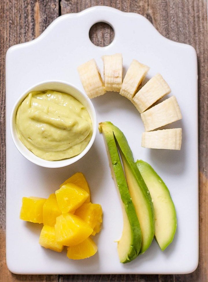 Image is of a white cutting board with a spread of produce and a small bowl filled with a creamy homemade baby food puree.