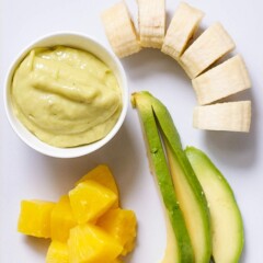 Image is of a white cutting board with a spread of produce and a small bowl filled with a creamy homemade baby food puree.