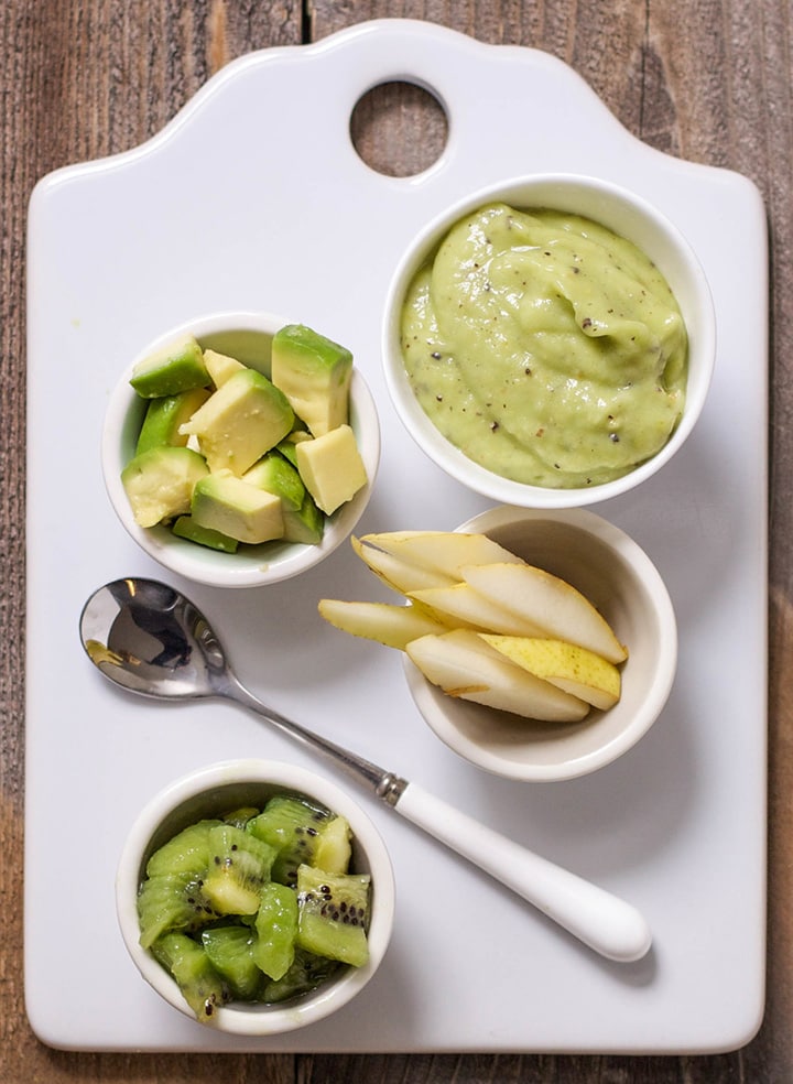 Image of a white cutting board on a wooden backdrop. On the board are several small bowls filled with produce - avocado, pears, kiwi and a small bowl with the smooth and creamy baby food puree.