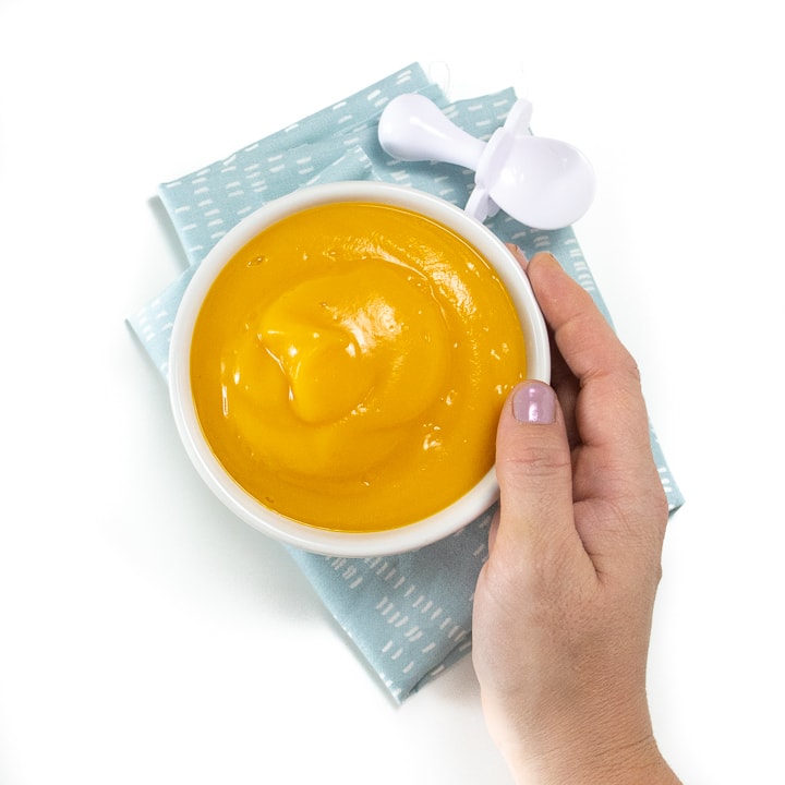 Small bowl of butternut squash puree along with a blue napkin and white baby spoon with a hand holding the bowl.