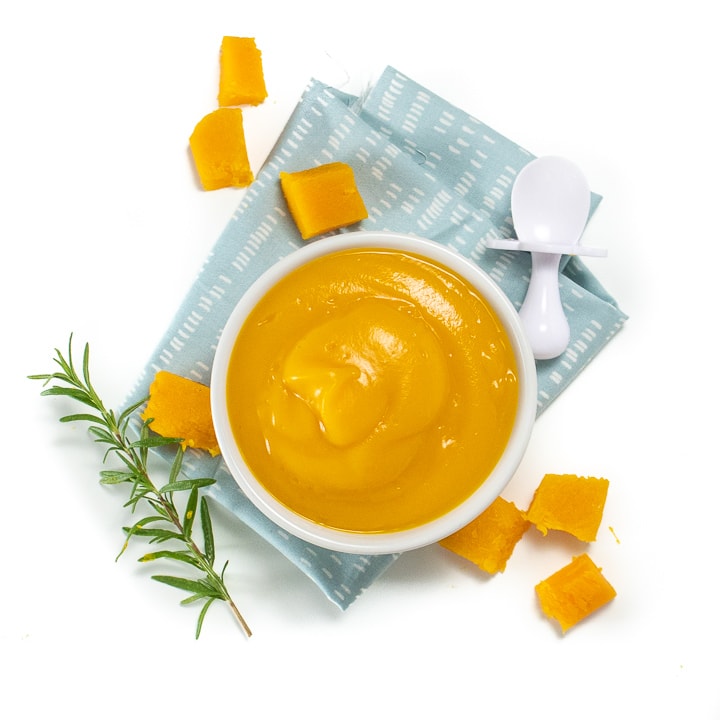 Spread of a small bowl of butternut squash puree along with chunks of butternut squash, rosemary and a white baby spoon.