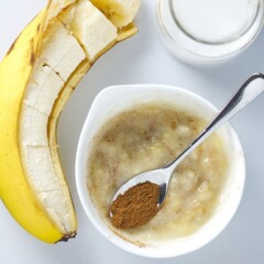 small round bowl filled with smashed baby food puree with a silver spoon resting inside the bowl filled with cinnamon. On a white background is a banana cut into chunks and small glass of coconut milk.