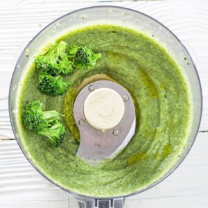 food processor is filled with a thick green broccoli puree with a few chunks of broccoli on the side.