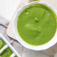 white bowl filled with green asparagus mint baby food puree. Bowl is sitting on a cutting board on a wooden white board.