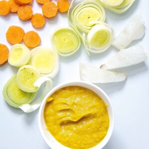 White cutting board with a spread of carrots, leeks and fish and a small bowl of baby food puree.