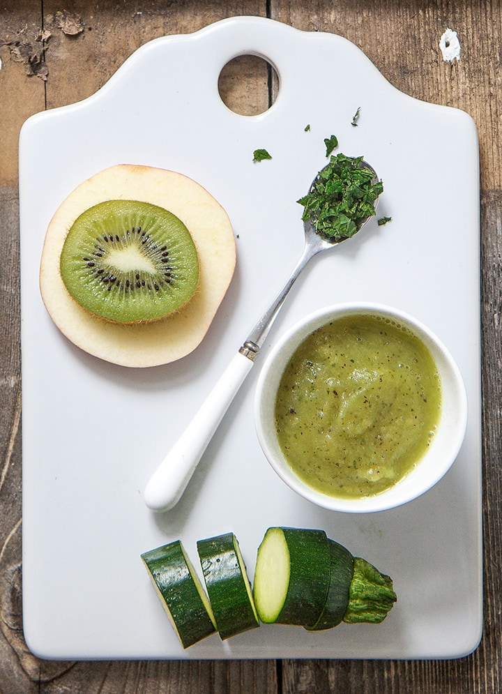 White cutting board with a spread of ingredients for this baby food puree - zucchini, apple, kiwi, mint and a bowl filled of the puree.