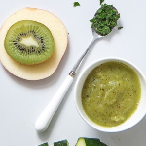 White cutting board with a spread of ingredients for this baby food puree - zucchini, apple, kiwi, mint and a bowl filled of the puree.