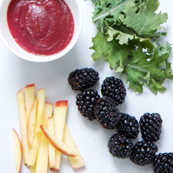 A bowl of fruit kale puree surrounded by blackberries, apples, and kale leaves.