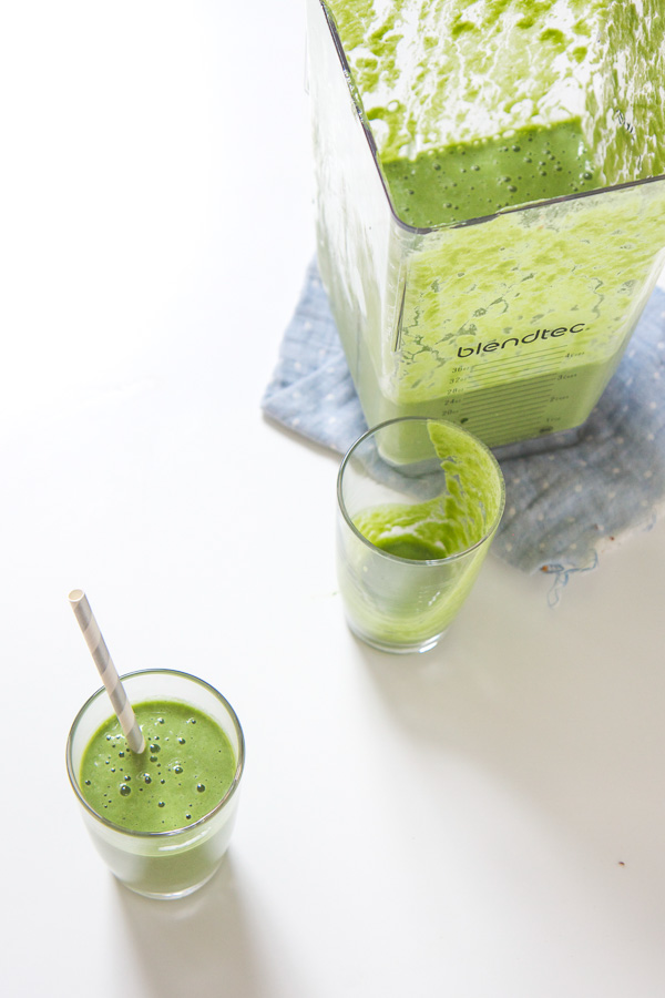 Two glasses of toddler approved green smoothies on a white surface with a blender of green smoothie.