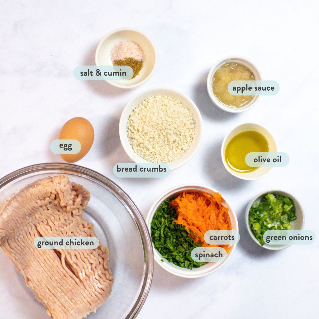 White kitchen counter with ingredients for chicken meatballs with graphics labeling each one - ground chicken, carrots, spinach, panko breadcrumb, eggs, applesauce, etc.