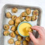 Metal baking tray on my counter, with a hand dipping a meatball into a yellow dip.