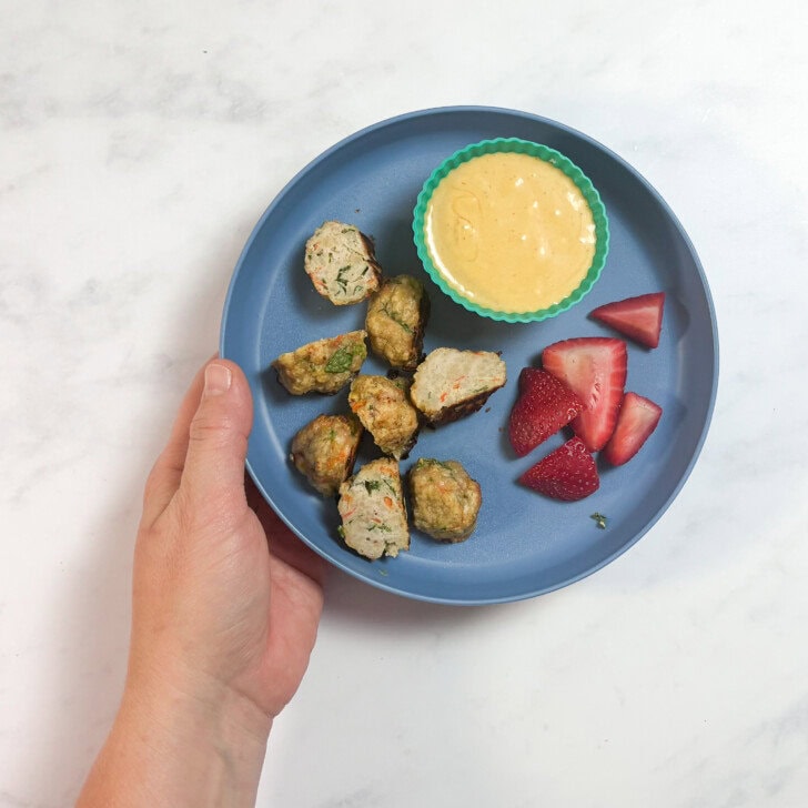 Hand holding a kids blue plate over my kitchen counter with how I serve these meatballs to my kids - meatballs cut in half with a dipping sauce and sliced strawberries.