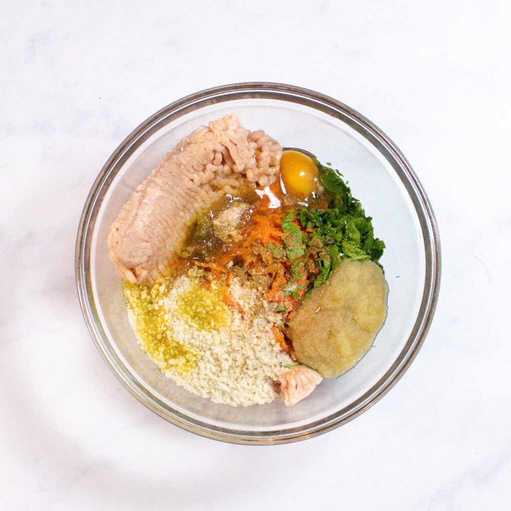 Glass bowl sitting on my white marble counterop, with ingredients for chicken meatballs inside.