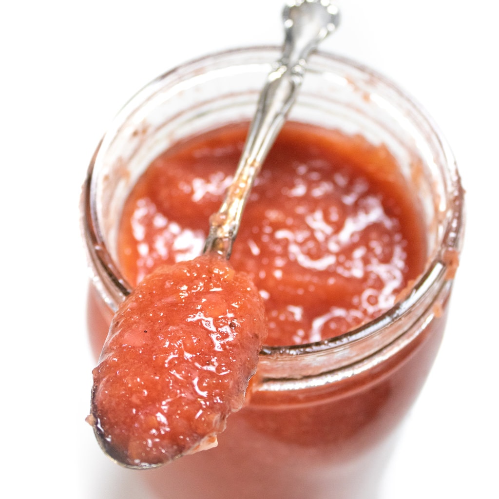 A jar full of strawberry applesauce with a silver spoon on top filled with strawberry applesauce.