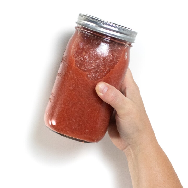 A jar of strawberry applesauce being held by hand against a white background.