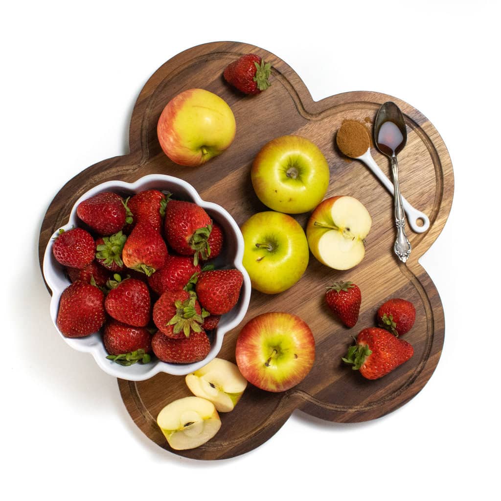A wooden cutting board with apples, strawberries, cinnamon and vanilla extract on top against a white background.