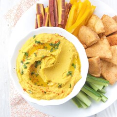 Carrot hummus in a white bowl with pita chips and vegetables.