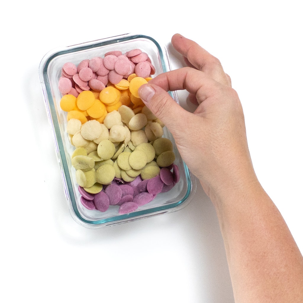 A rectangle glass container full with five different colors of yogurt melts for baby with a hand picking up an orange circle.