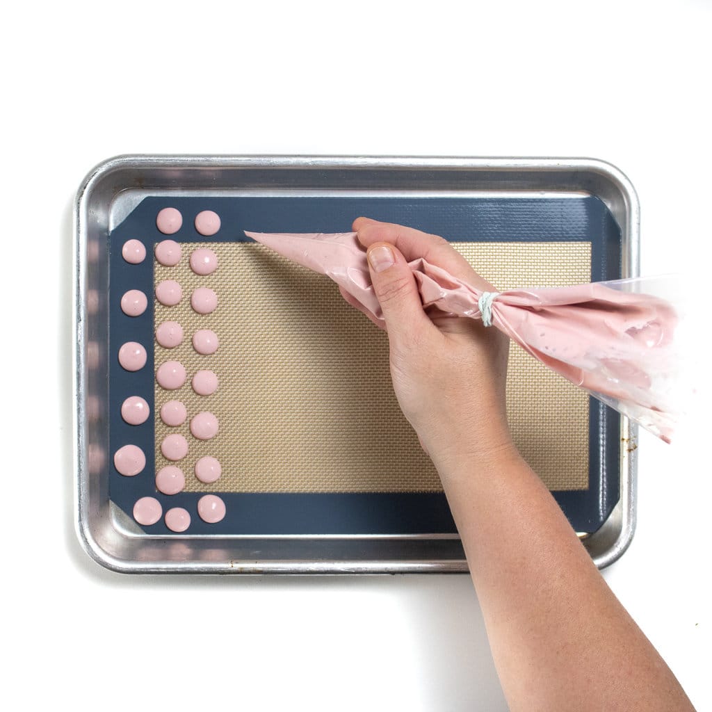 A hand piping pink yogurt melts onto a silver baking sheet.