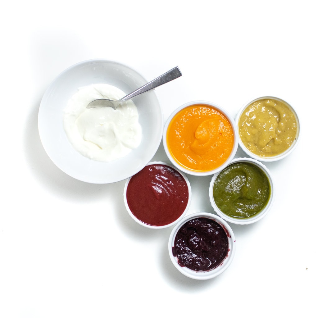 Bowl of yogurt is surrounded by five different colors of baby food purée against a white background.