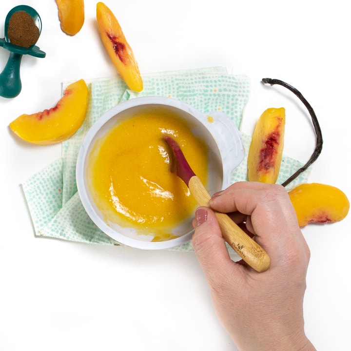 Small gray bowl filled with a peach puree for baby with peaches and vanilla bean besides it and hand reaching into the frame to stir the puree.