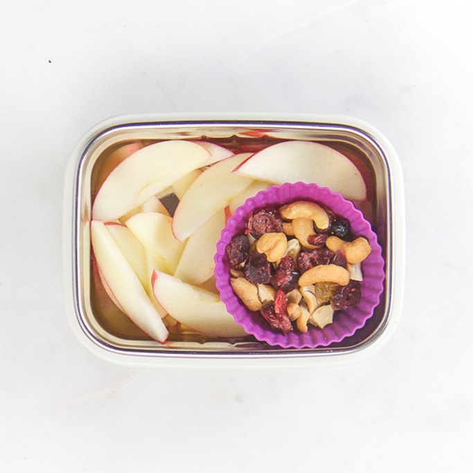 Rectangle kids bento box with healthy snacks for kids - apple slices with a purple muffin mold filled with trail mix