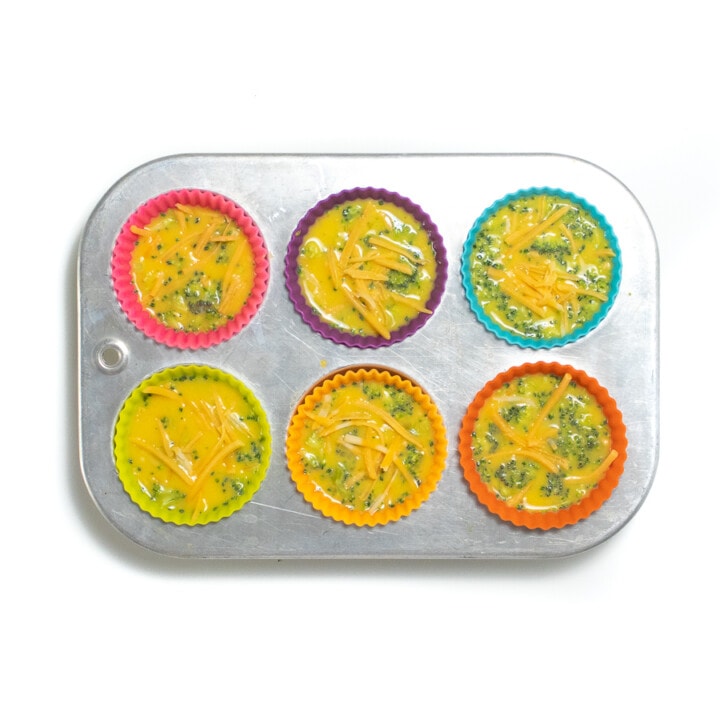 Silver muffin tin with colorful silicone liners filled with broccoli cheese egg cups.
