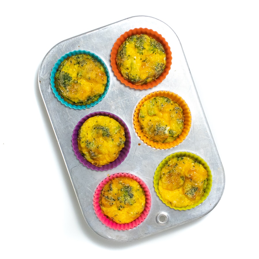 Hands holding a 6-cup muffin tin filled with colorful liners and broccoli egg cups.