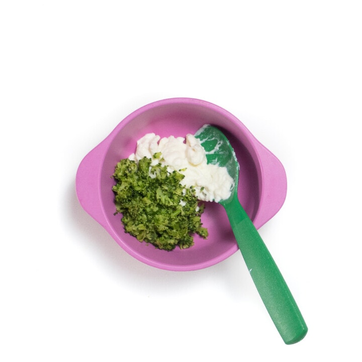Pink baby bowl with a green spoon and a spoonful of ricotta and chopped steamed broccoli.
