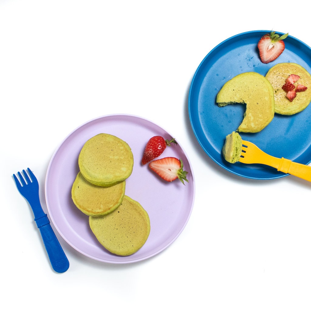 A purple and blue toddler play with spinach pancakes chopped strawberries and a orange fork against a white background.