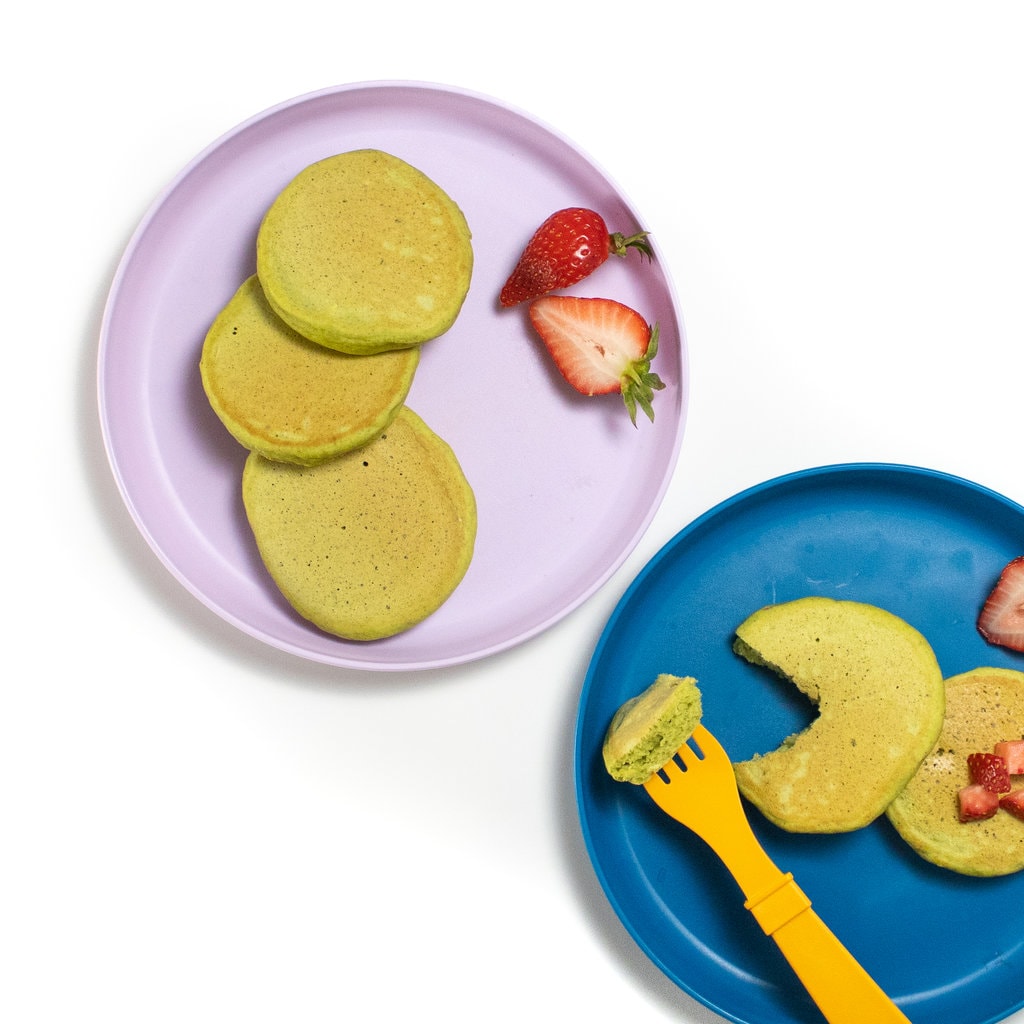 A purple and blue toddler play with spinach pancakes chopped strawberries and a orange fork against a white background.