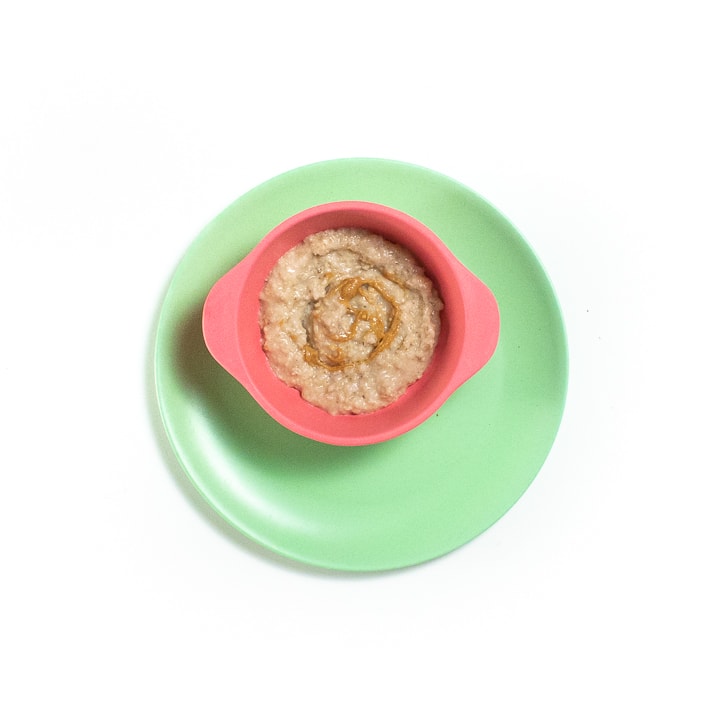 Pink baby bowl on a green plate. In the bowl is oat cereal and a swirl of peanut butter.