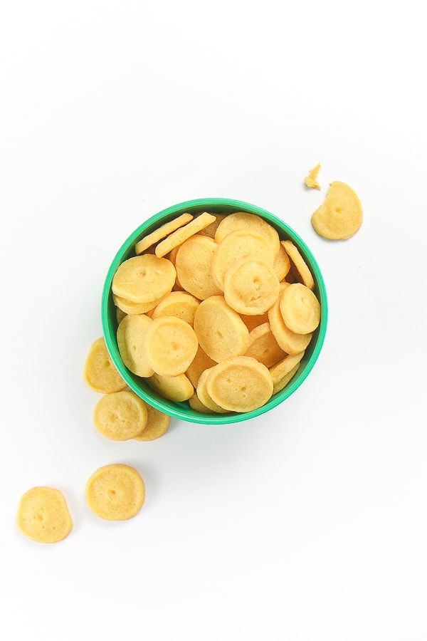 small green plastic bowl filled with yellow cheddar crackers. Some crackers on spilled out on a white background.