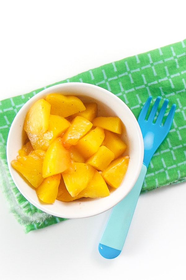 baby-led weaning snack - White bowl filled with peach chunks. Sits on a green napkin and blue fork is nestled into bowl. 
