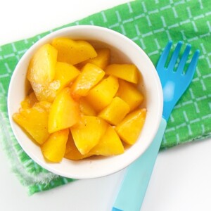 baby-led weaning warm peaches finger food for baby or toddler - small white bowl on top of green napkin with a blue fork. Inside of bowl are chunks of peaches with a sprinkle of nutmeg.